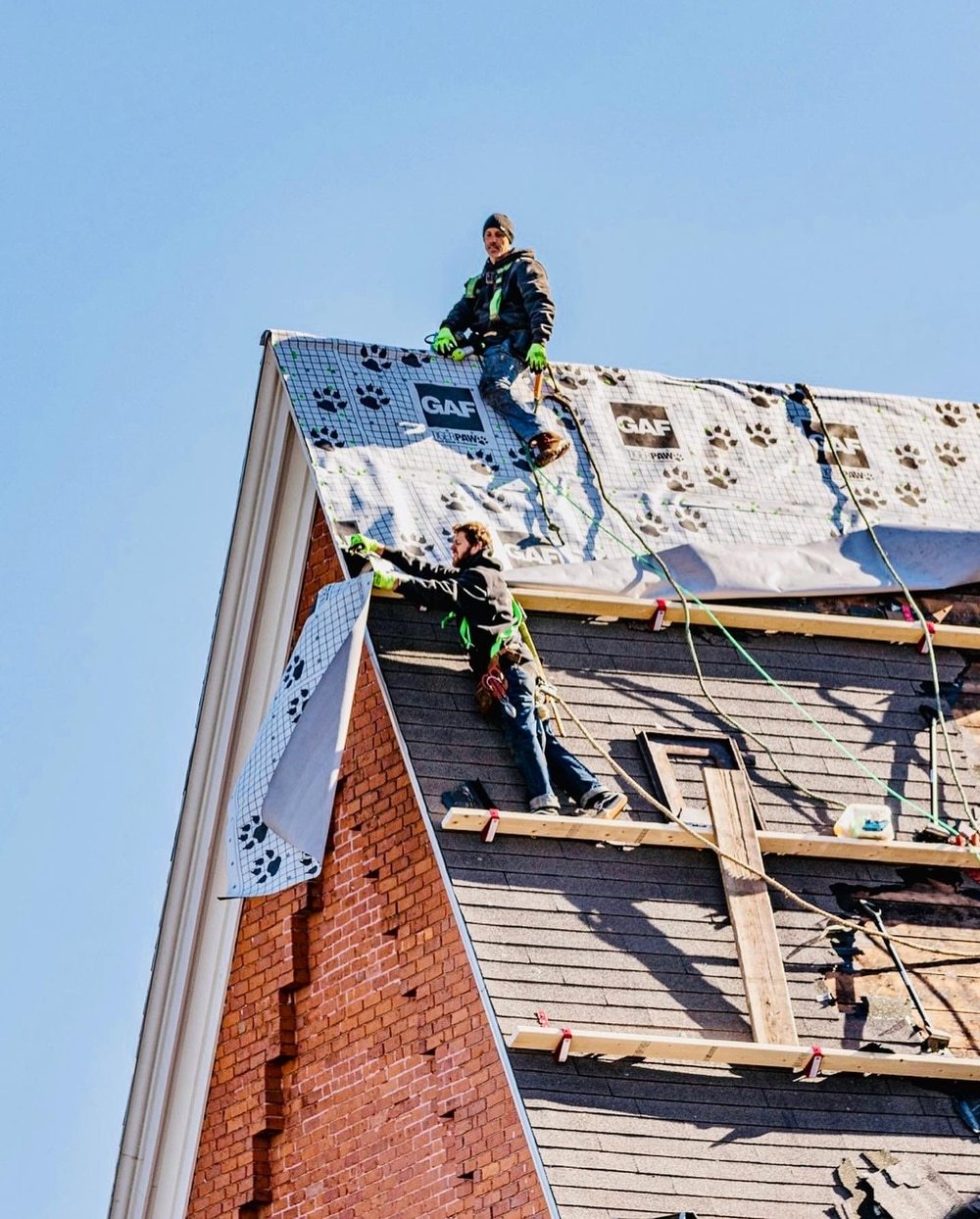 Chris Battaini Roofing - Two workers in safety gear repair the steep roof of a brick building, using ropes and ladders, under a clear blue sky.