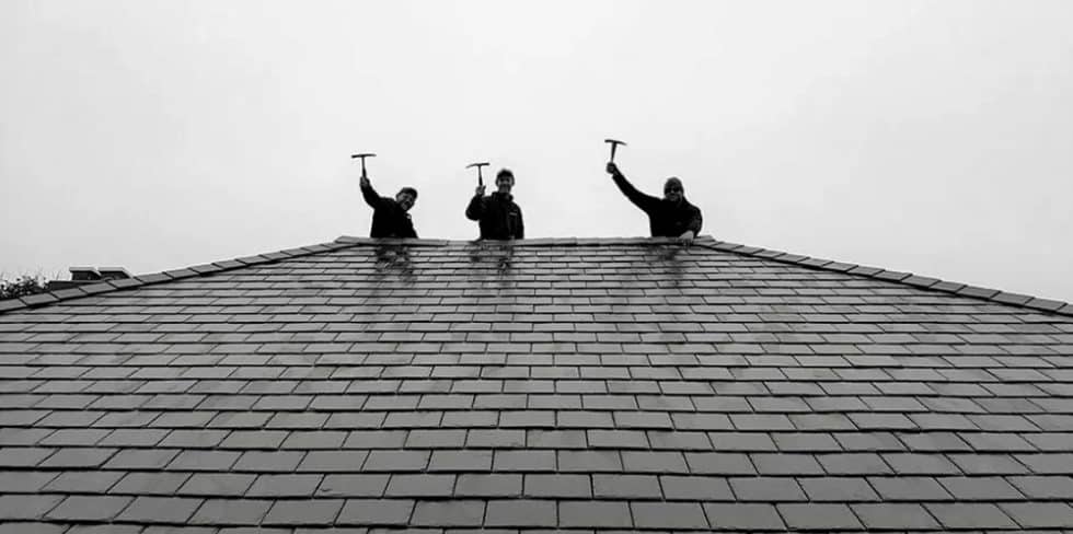 Chris Battaini Roofing - Three people stand at the top of a tiled roof, each holding a hammer raised in the air against a cloudy sky.