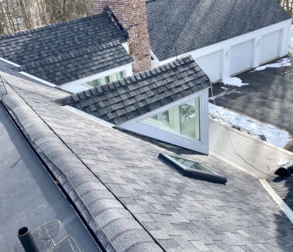 Chris Battaini Roofing - View from a rooftop showing gray asphalt shingles, two dormer windows, a chimney, garage doors, and patches of snow on the ground.