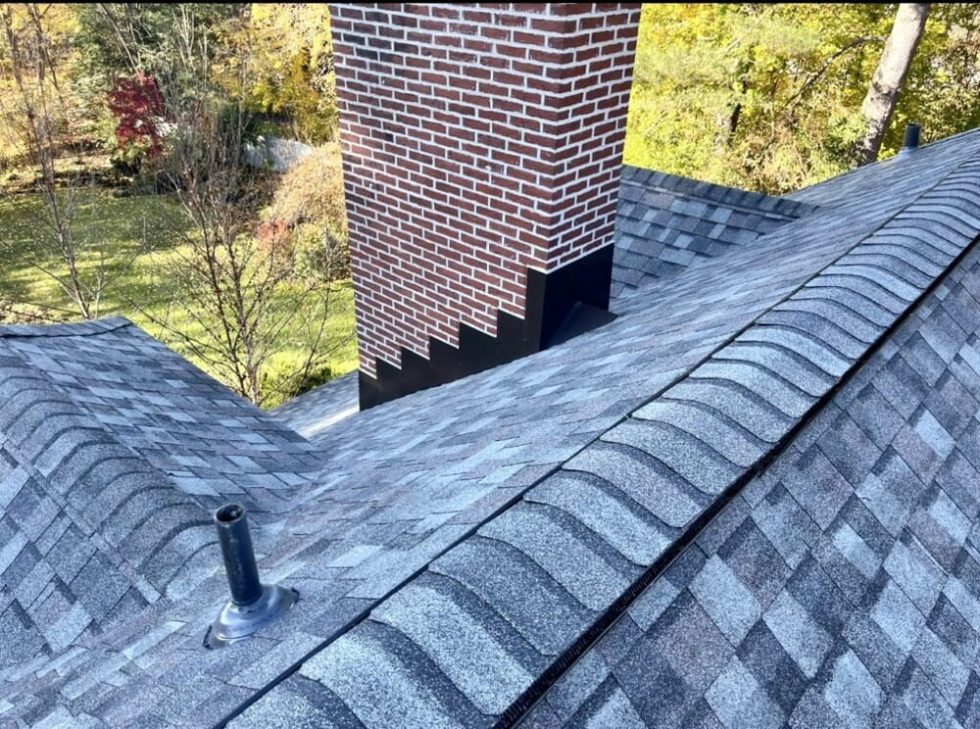 Chris Battaini Roofing - Gray shingle roof with a brick chimney and a vent pipe, surrounded by trees with autumn foliage in the background.