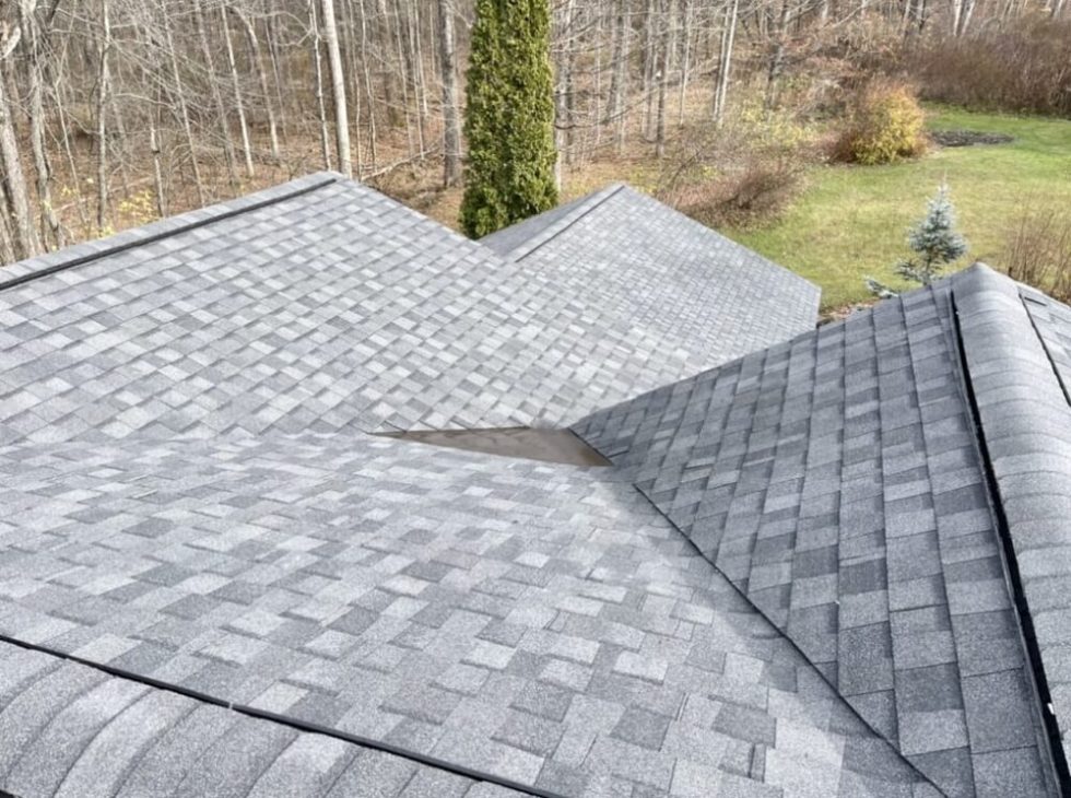 Chris Battaini Roofing - Gray shingled roof with intersecting ridges and valleys, surrounded by trees and a grassy yard with some shrubs in the background.