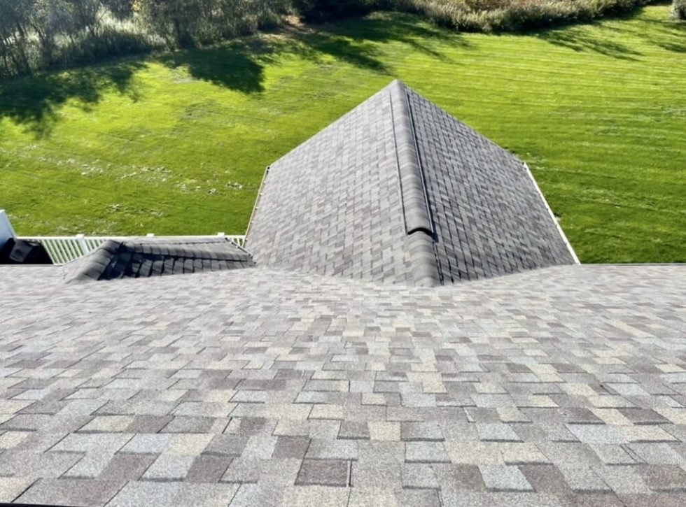Chris Battaini Roofing - View from a rooftop with gray shingles, overlooking a green, well-manicured lawn and trees in the background.