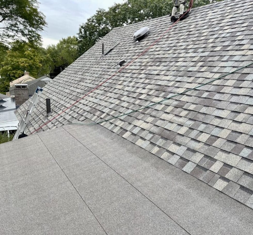 Chris Battaini Roofing - Gray shingles cover a sloped house roof, with safety ropes and equipment visible; trees and part of another house are in the background.
