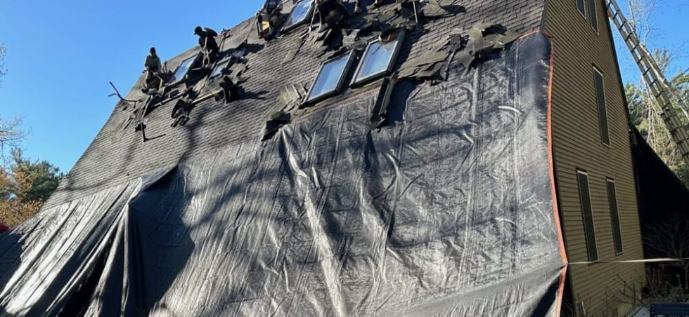Chris Battaini Roofing - Workers repair a steep roof covered in tarps and debris, with ladders propped against the house under a clear blue sky.