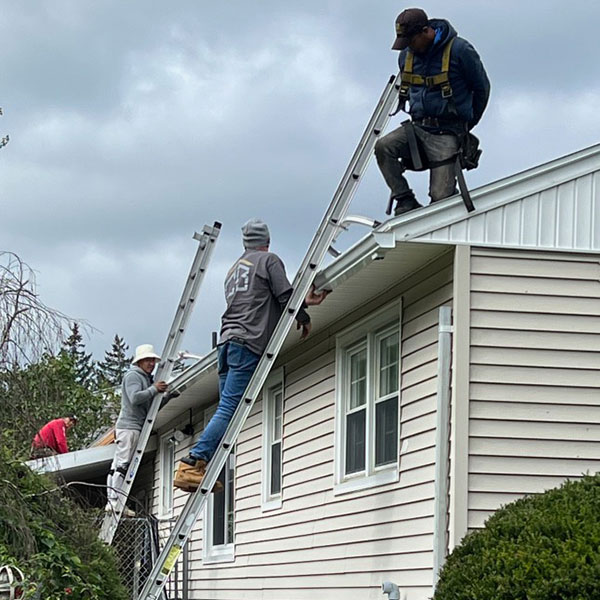 Chris Battaini Roofing - Four workers on ladders and a roof install or repair gutters on a beige house under a cloudy sky.